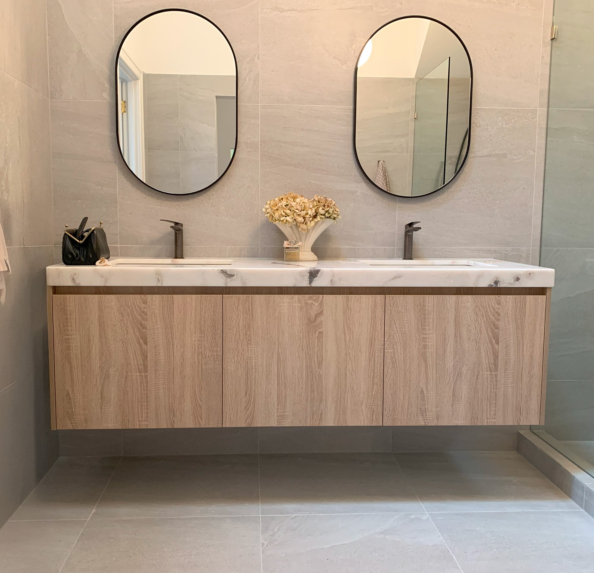 Floating timber-look double vanity with marble top, two oval mirrors and tiled floor